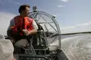 An airboat pilot at the controls with the fan cage behind him