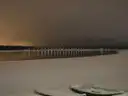 Kayaks pulled up on a beach at night, with lights along a pier