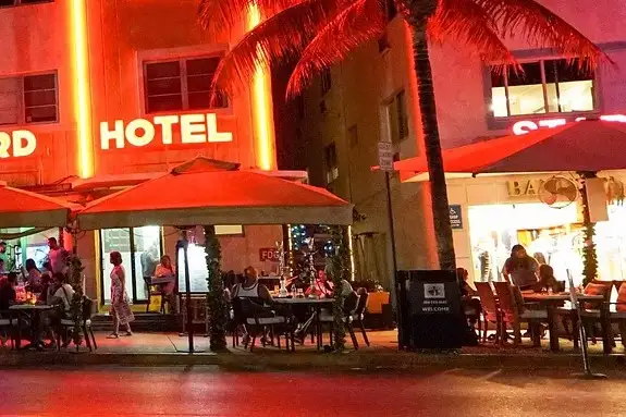 People at the outdoor tables of a neon-lit Ocean Drive hotel at night, with a woman walking past